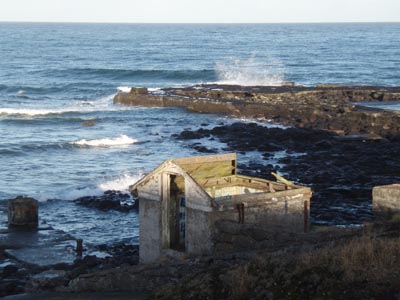 Photo of abandoned pump house with missing roof near rocky shore.